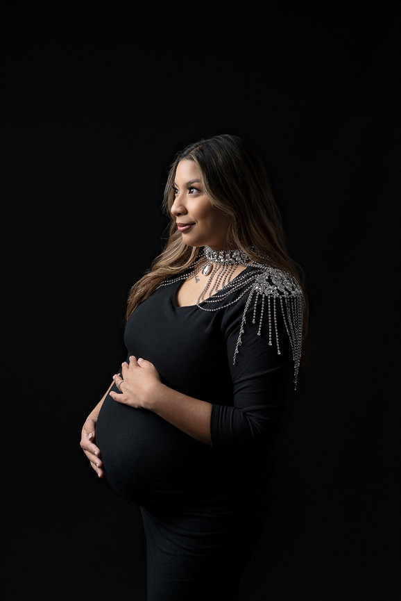A pregnant woman in a black dress stands against a dark background, cradling her belly. She wears a decorative shoulder piece with silver embellishments, looking thoughtfully to the side.