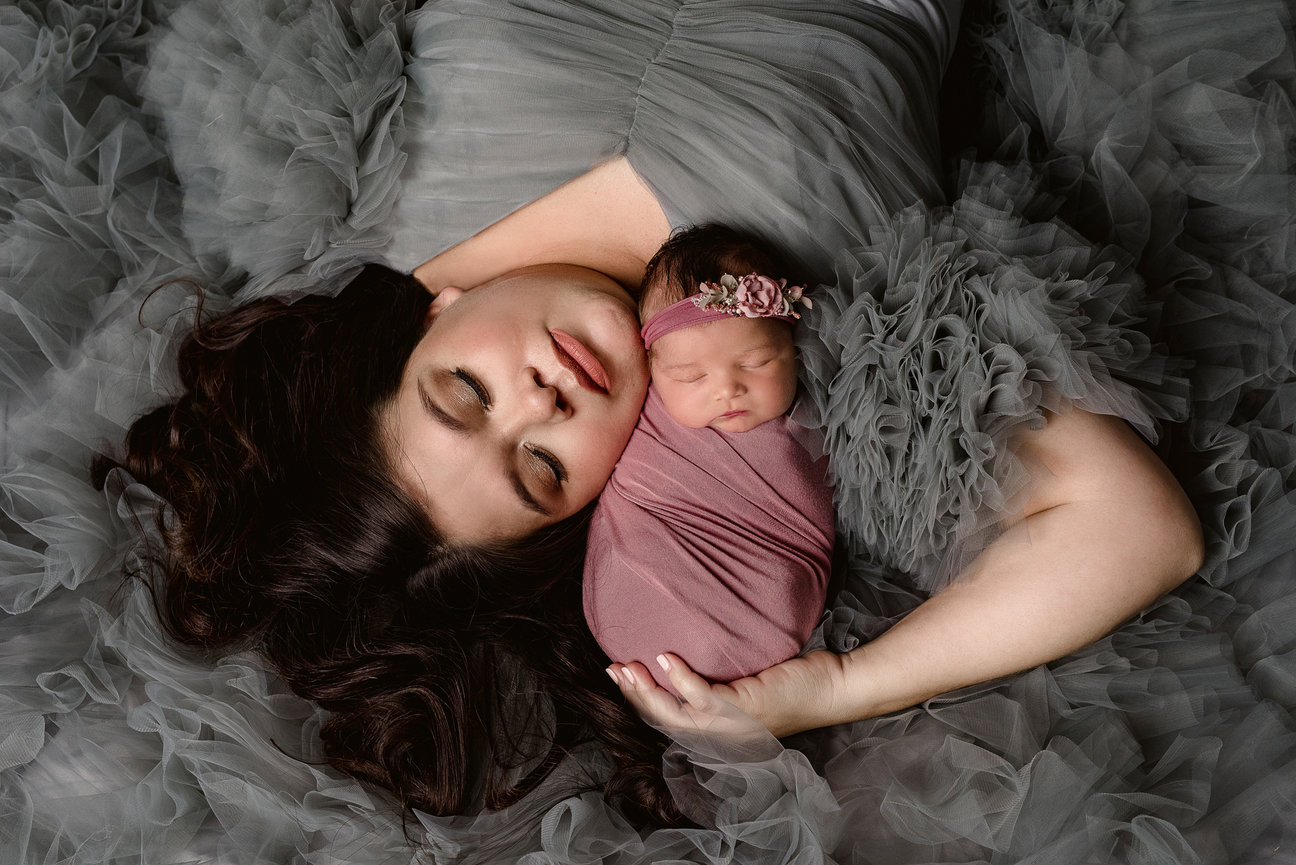 In Barrington Hills, a woman in a gray, ruffled gown lies peacefully with her eyes closed, holding a sleeping baby wrapped in a mauve swaddle and wearing a floral headband. They are both surrounded by soft, flowing fabric.