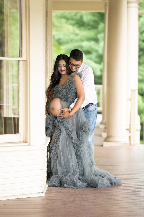 The image captures a smiling couple standing on a porch. The woman, in a flowing gray gown, lovingly cradles her pregnant belly, while the man, dressed in a white shirt and jeans, embraces her from behind. They stand against the backdrop of columns.