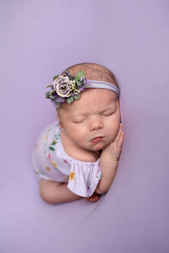 In Lakewood, a sleeping baby wearing a floral headband and a white outfit with tiny flower patterns peacefully rests against a soft purple backdrop. The babys hand gently supports their cheek.