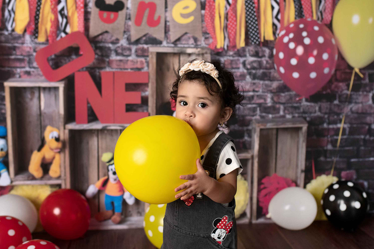 A toddler celebrates their 1st birthday, holding a yellow balloon in front of a ONE themed backdrop adorned with colorful decorations, polka dot balloons, and plush toys. The child sports overalls featuring cartoon characters and a floral headband.