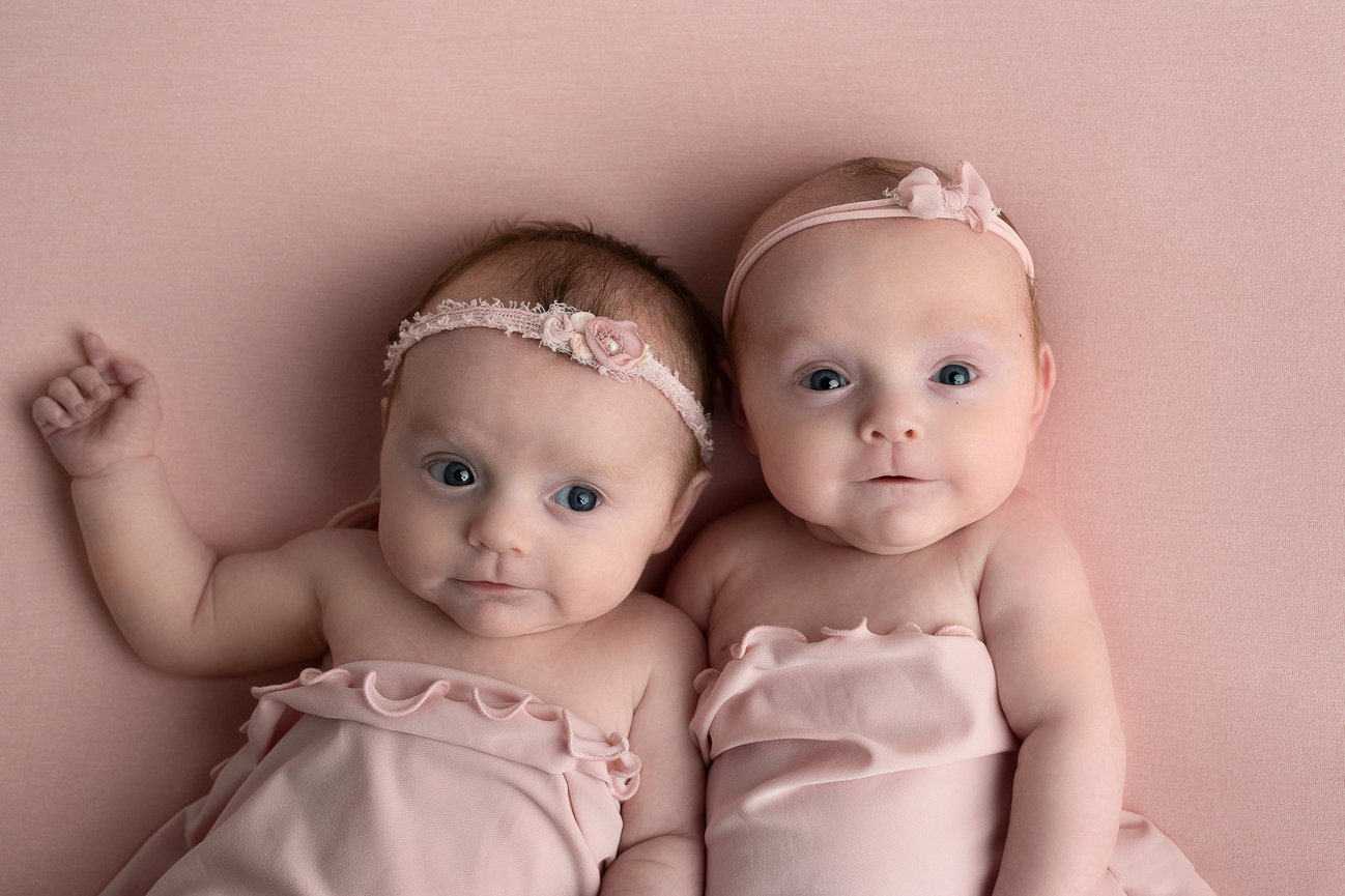 Two babies lie on a soft pink surface in Sleepy Hollow, wrapped in pink fabric with matching headbands adorned with flowers. They look directly at the camera, with one baby slightly raising their arm.