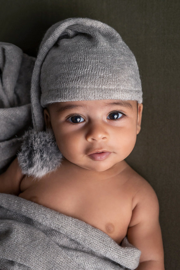 A baby wearing a gray knitted hat with a fluffy pom-pom lies on a matching gray blanket, ready for the best holiday portraits. The baby has a calm expression, looking directly at the camera against a soft, muted background.