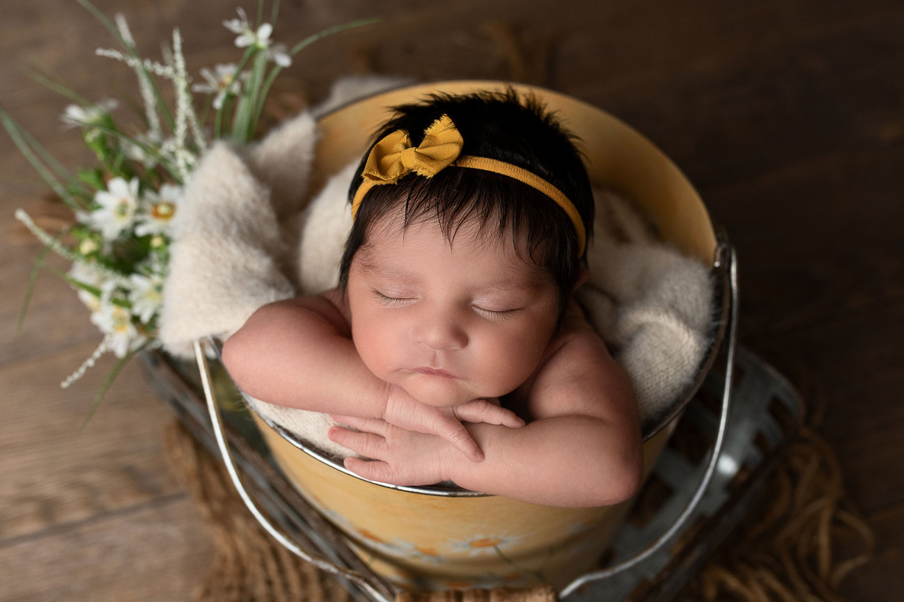A sleeping baby with dark hair rests inside a yellow metal bucket, wrapped in a soft cream blanket. The baby, adorned with a yellow headband and bow, lies peacefully as if dreaming of West Dundee. Nearby, small white flowers on a wooden surface.