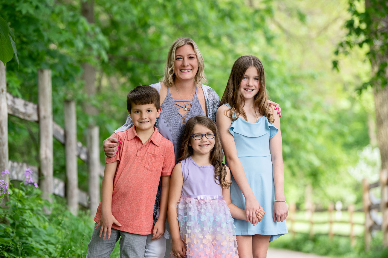 A smiling woman stands outdoors on a path lined with greenery, captured by an Elgin family photographer. She poses with three children: one boy in a peach shirt and two girls in pastel dresses. They look happy under the soft, natural lighting.