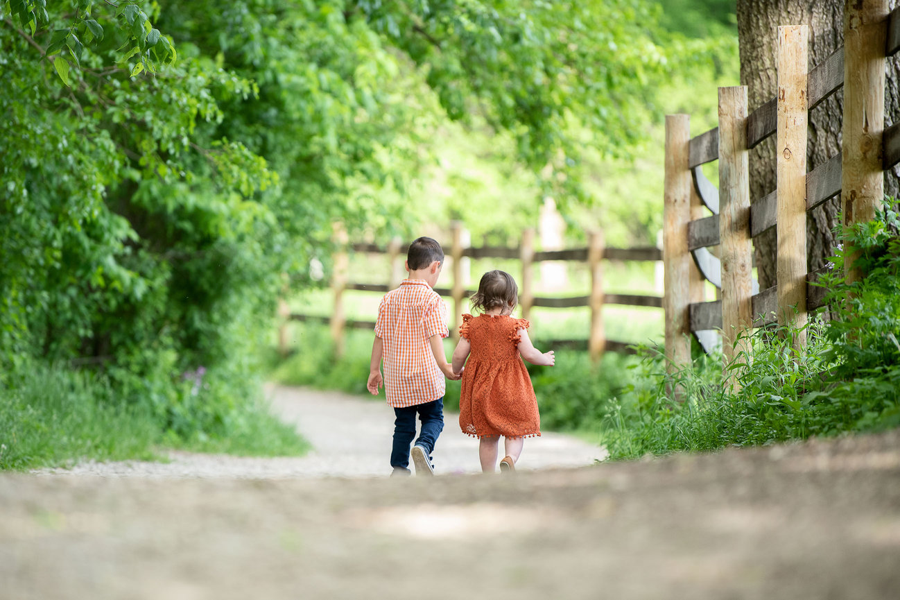 A young boy and a little girl, both in matching orange outfits, stroll hand-in-hand along a dirt path surrounded by lush green trees and a wooden fence under the sun. This charming scene captures the essence of Illinois in the town of Algonquin.
