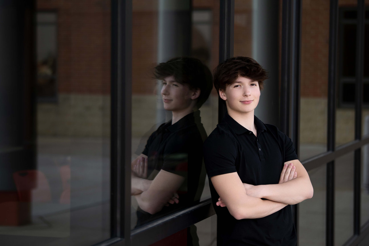 A young person with short dark hair in a black shirt stands confidently with arms crossed, leaning against a glass window that reflects their image like a captured moment from graduation photos. The textured brick and stone wall in the background.