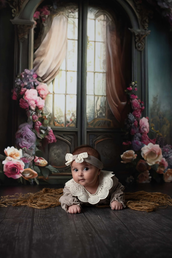 A baby wearing a lace-trimmed outfit and bow headband lies on a dark wooden floor in Streamwood, Illinois. The backdrop features ornate, vintage-style windows adorned with curtains and surrounded by an array of colorful, blooming roses.