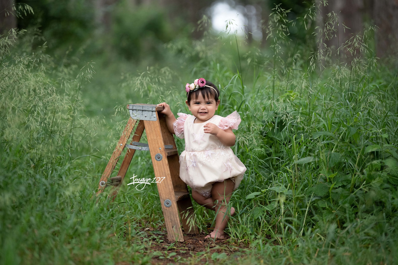 A baby in a light pink outfit with a floral headband stands beside a small wooden ladder in a lush, green field just outside of Chicago. Smiling amid the tall grass and trees, the scene captures a serene setting reminiscent of an urban escape.