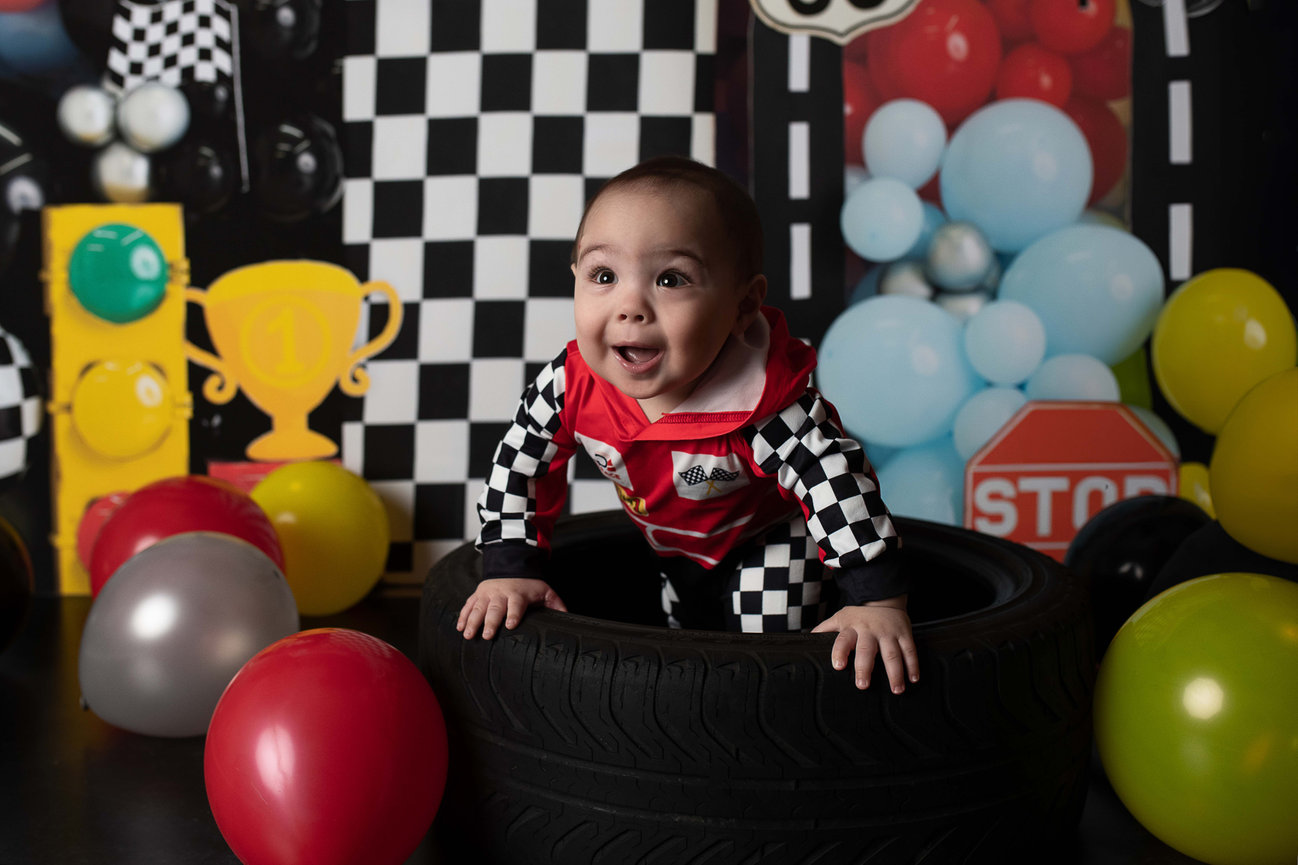 A smiling baby in a racetrack-themed outfit sits inside a tire surrounded by colorful balloons. The background features a checkered pattern, a trophy, and a cartoonish traffic sign.