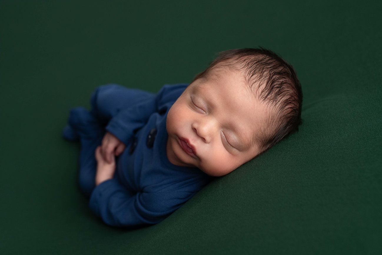 A newborn baby peacefully sleeps on a soft, dark green surface. The newborn is wearing a cozy blue outfit, with hands gently crossed over the chest. The serene scene captures the innocence and tranquility of infancy.