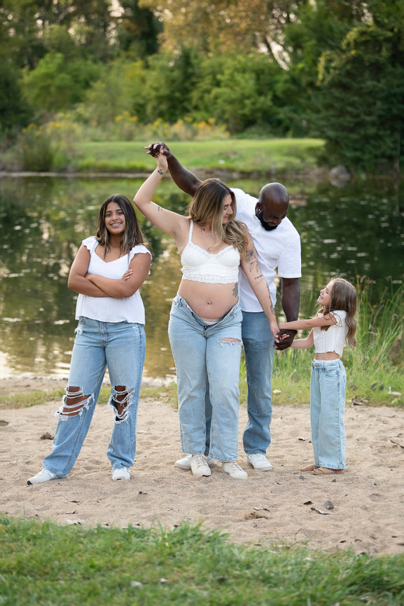 A family of four stands by a pond. The father is twirling the mother, who holds the hand of a young girl. The older girl stands to the side, smiling. They are all casually dressed in white tops and jeans. Trees are visible in the background.
