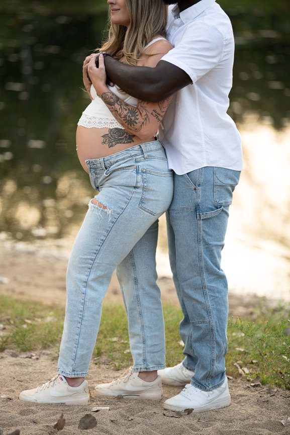 A pregnant woman with tattoos is embraced by a man by the tranquil Barrington lake. Dressed in casual clothing, including jeans and sneakers, they share an intimate moment surrounded by lush greenery and water, creating a serene backdrop.
