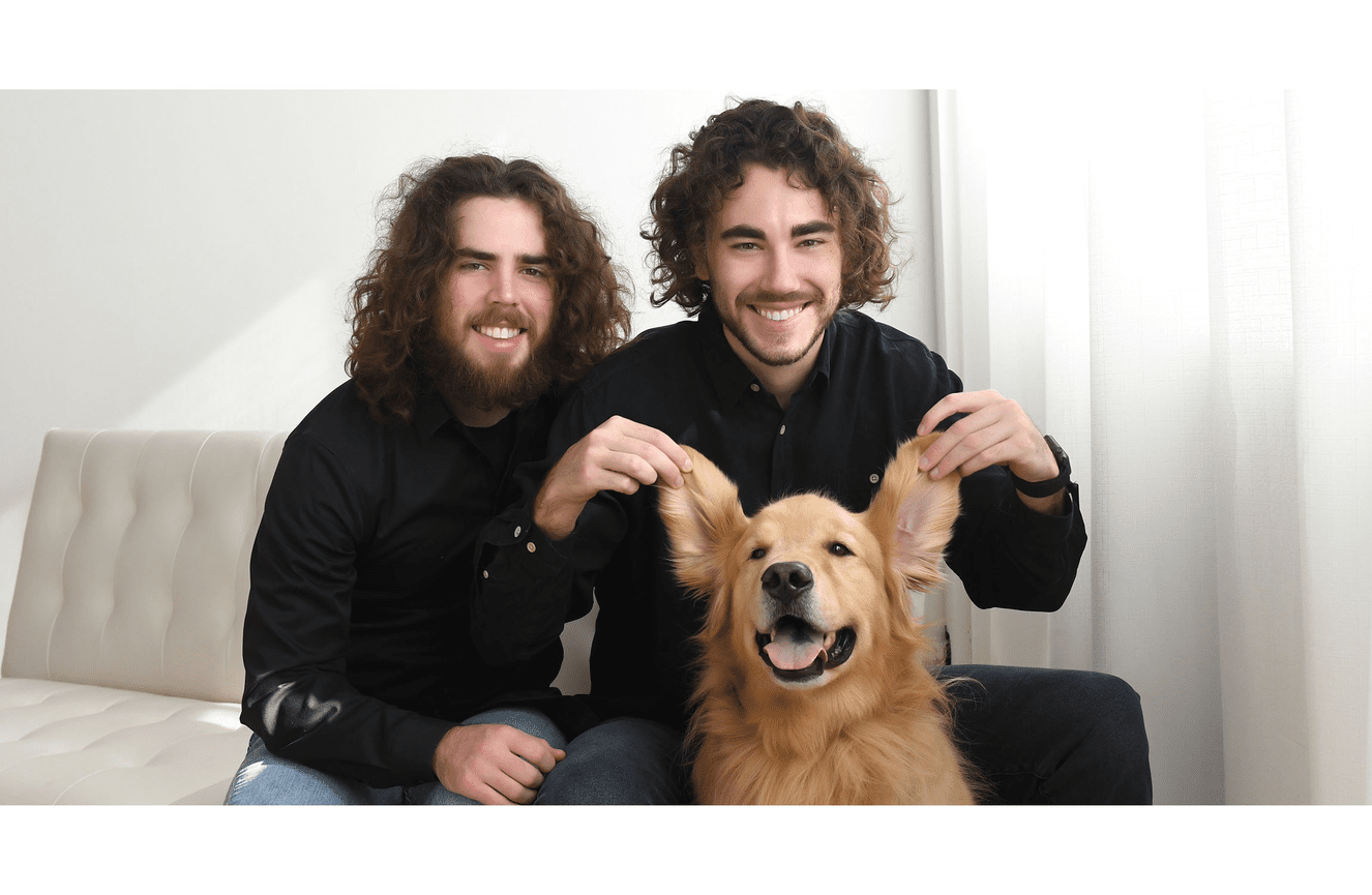 Two college aged brothers on a white couch holding up the ears of their smiling golden retriever dog in the Kliks Photography studio.