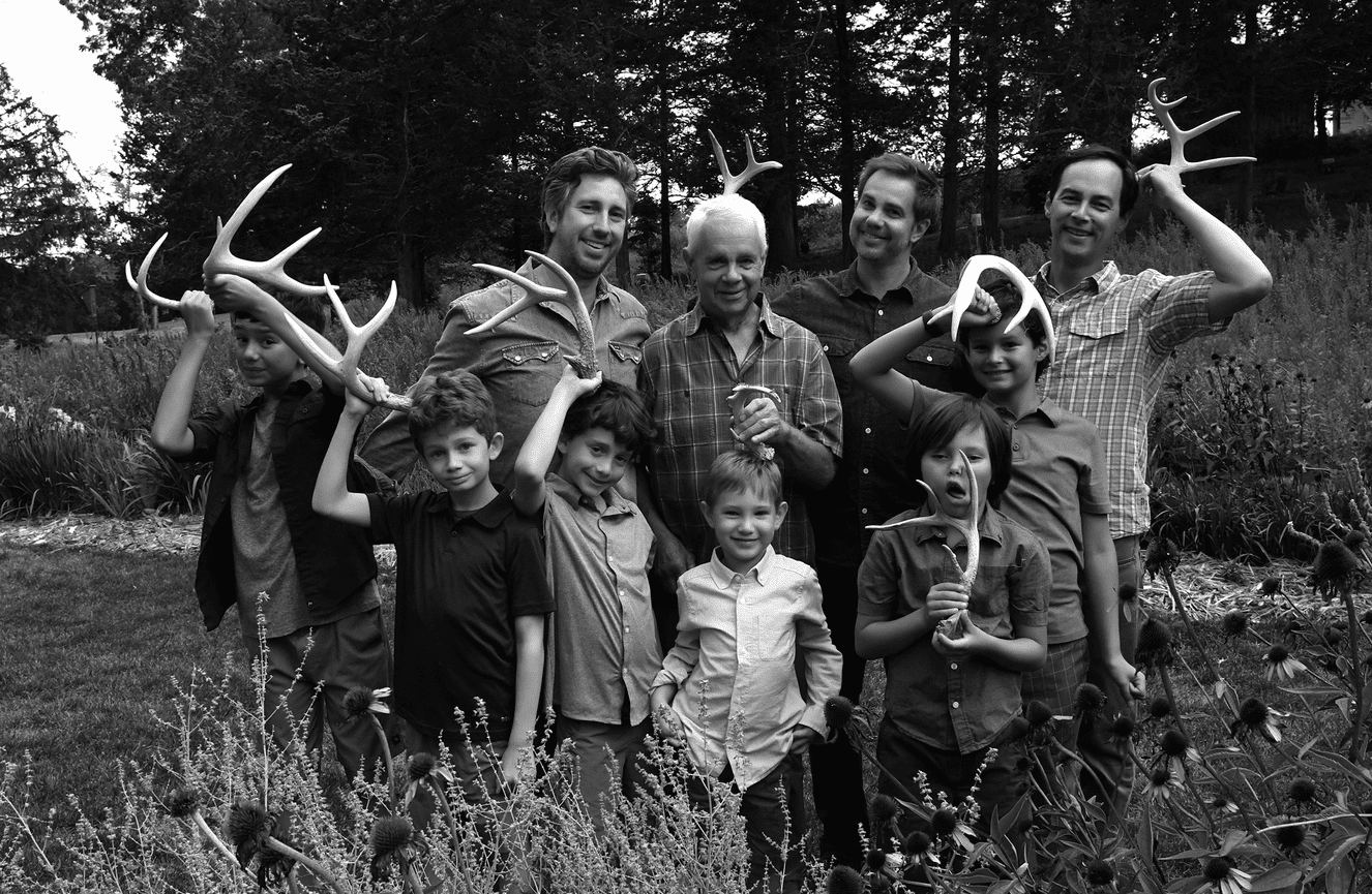 A black and white photo of three generations of men making funny faces with deer horns they found in the fields.