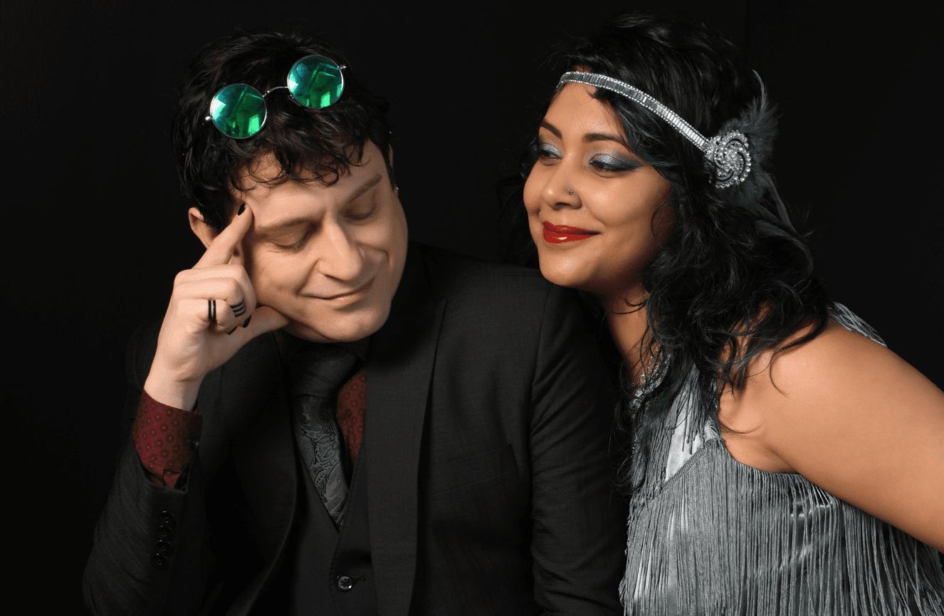 Joseph in a three-piece suit and mirrored sunglasses looks lovingly at his partner in her 1920's lapper dress during a photo shoot at Kliks Photography.