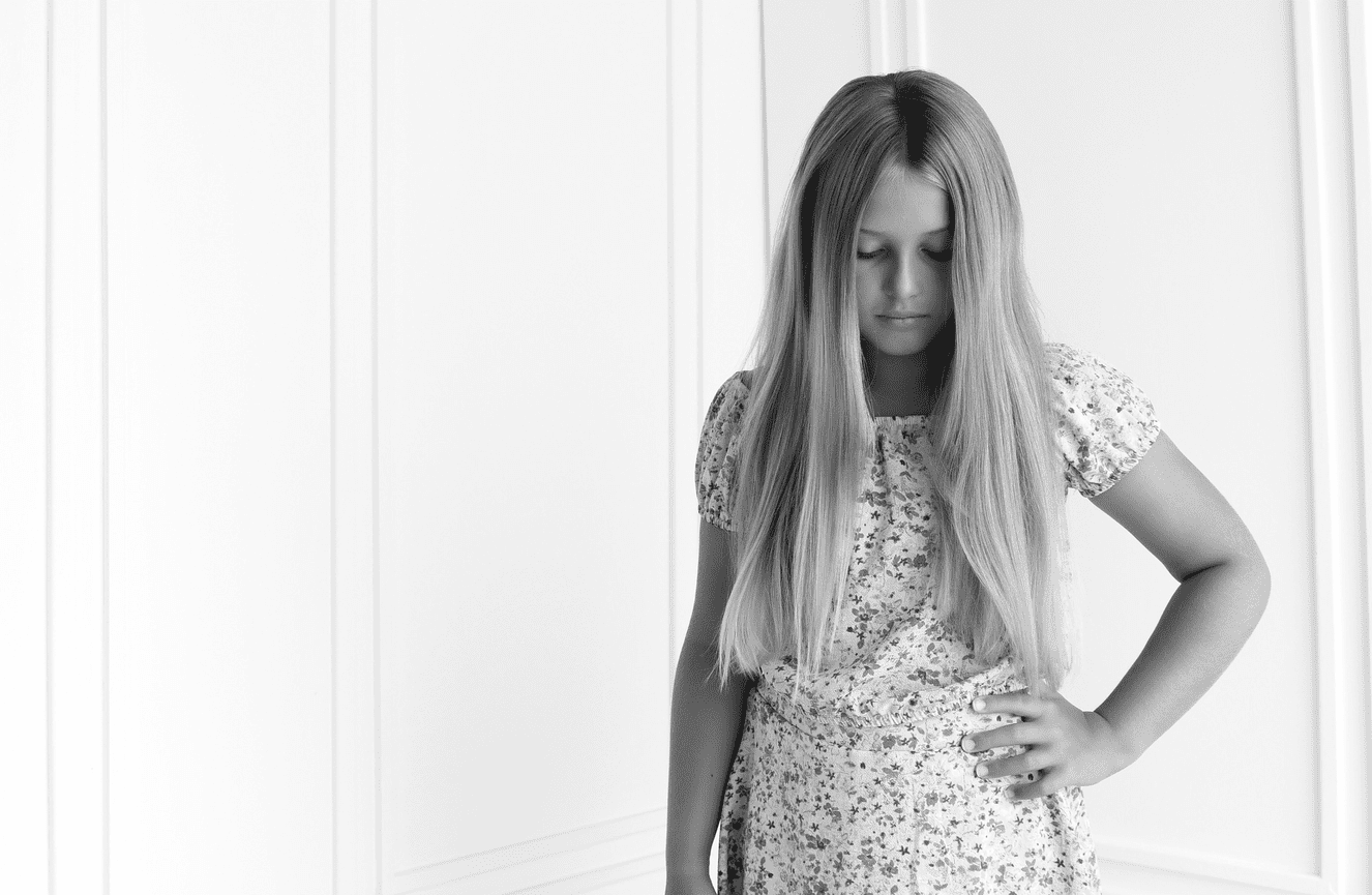 A somber pre-teen girl looking down with her long blonde hair nearly covering her face.