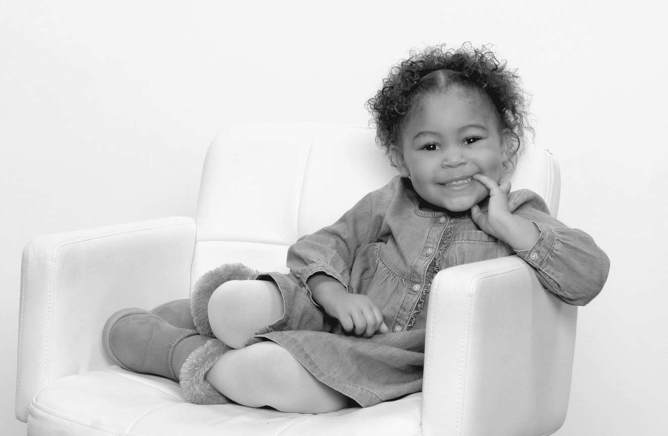 An African-American toddler girl in a jean dress and Ugg boots smiling during her photo session at Kliks.