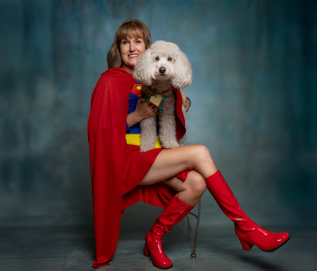 Smiling woman in a red superhero costume holding a fluffy white dog against a blue background.