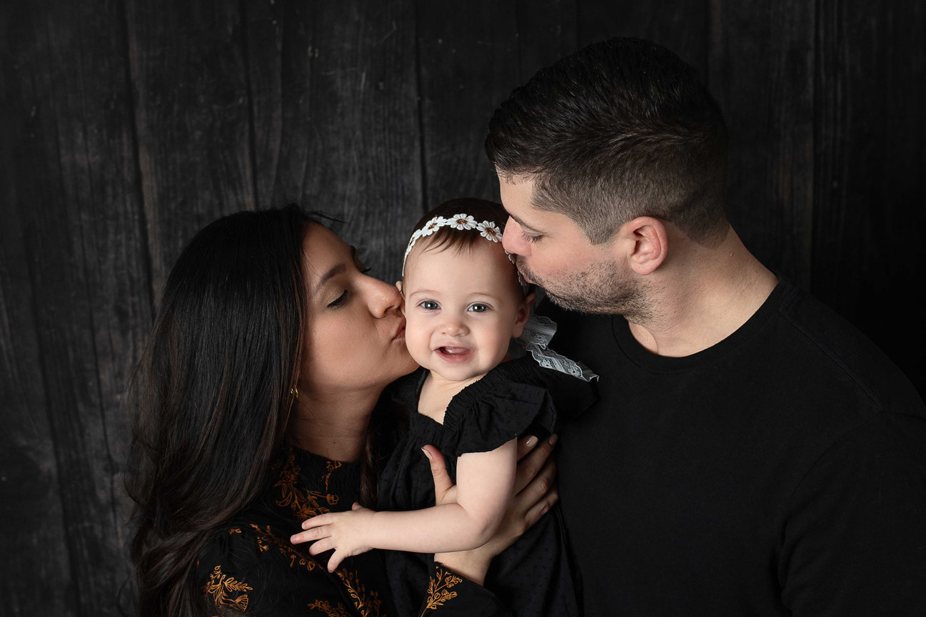 A couple kissing a baby wearing a black dress and headband, posed against a dark wooden background.
