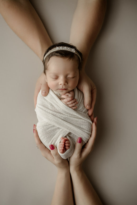 A newborn baby swaddled in white, held gently by two hands on a soft background.