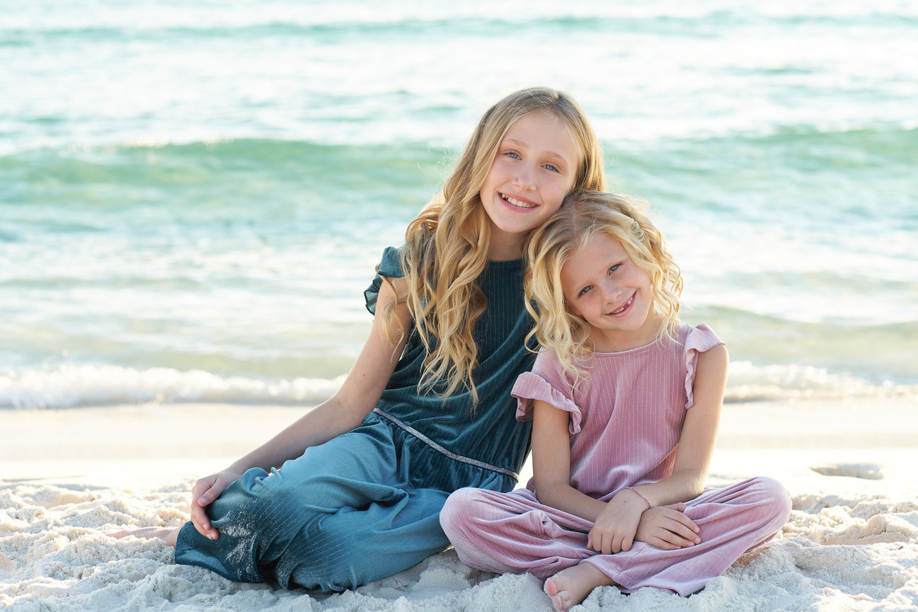 Two young girls with long hair sitting on a sandy beach with ocean waves in the background.