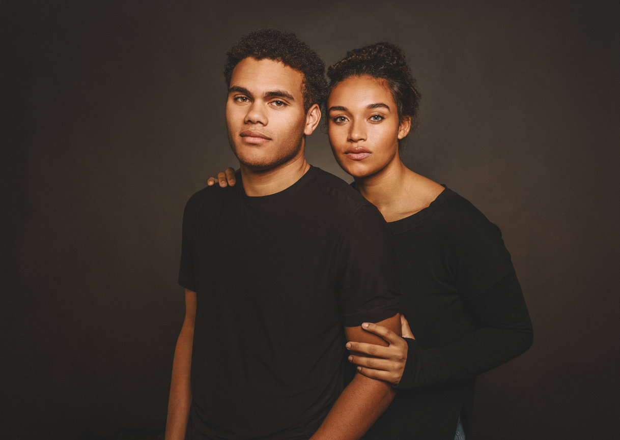 Studio portrait of two people in black shirts, one standing behind and embracing the other.