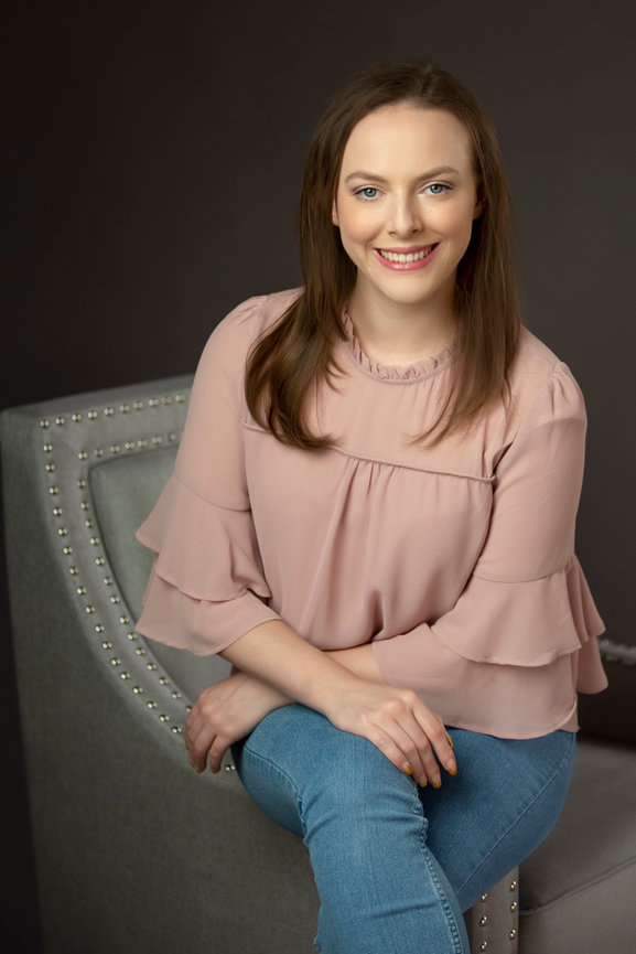 Modern business portrait head shot of a young woman seated on the arm af an elegant chair, with her legs crossed, arms resting on her knee with a happy and connected expression to the camera.