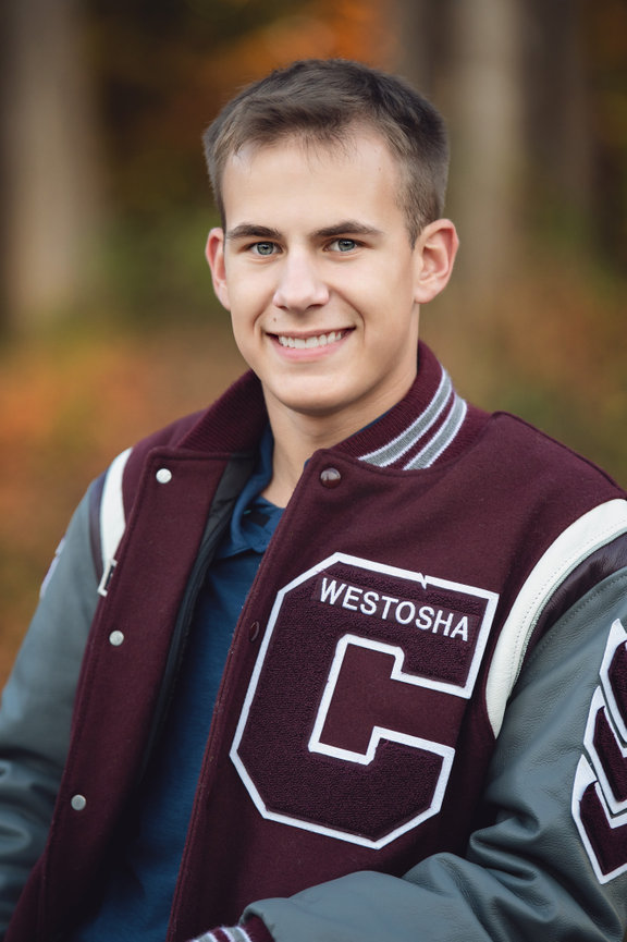 a high school senior guy wearing a maroon letterman jacket stands outdoors, smiling, with autumn foliage in the background at petrifying springs park.
