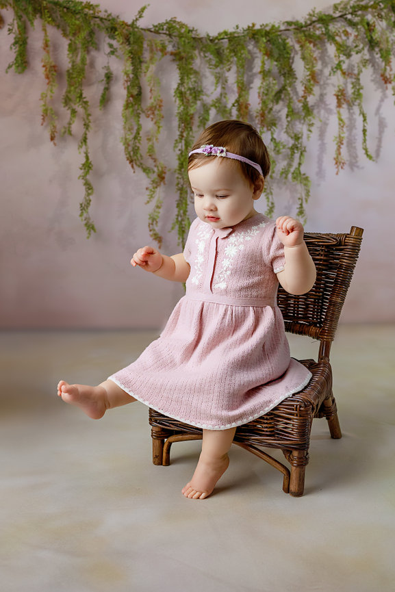 Baby girl in a pink dress sitting on a wicker chair with a floral backdrop in Kimberly A King Photography's Studio in Warsaw, VA