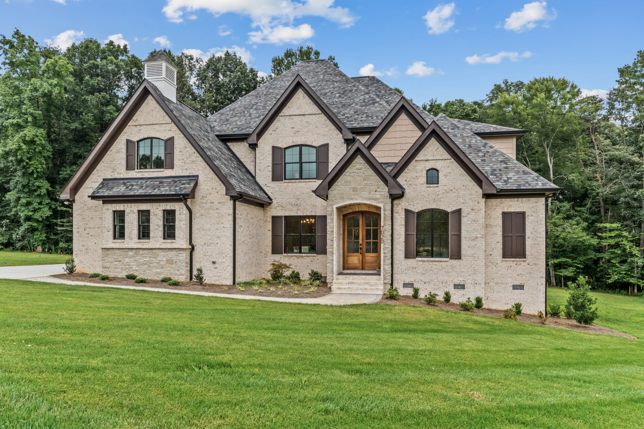 Large beige brick suburban house with gabled roof, green lawn, and trees under a blue sky