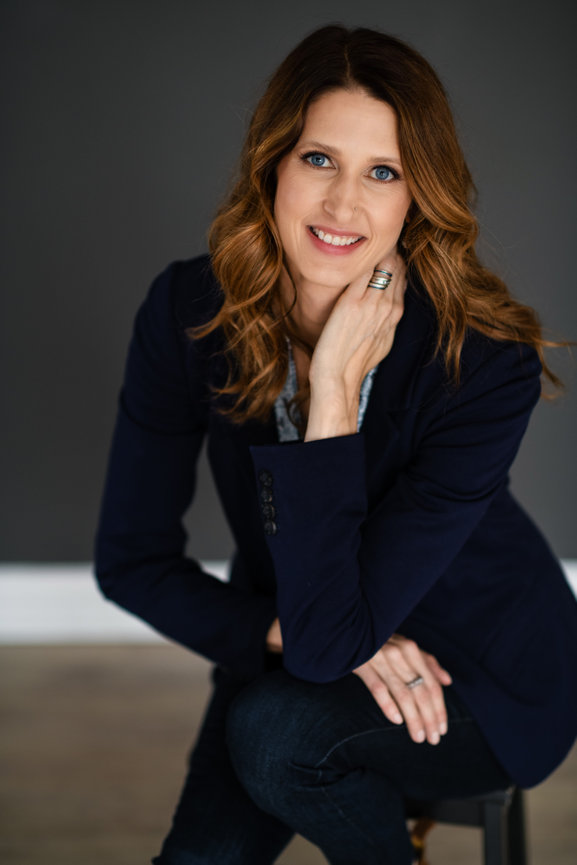 Professional portrait of a woman with wavy hair, wearing a navy blazer, smiling and posing thoughtfully against a dark background. Dublin Ohio Headshots
