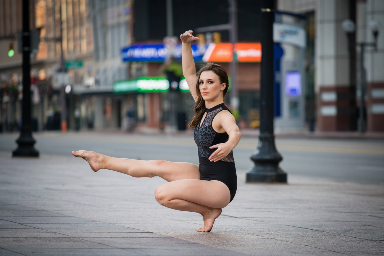 Dancer in a black leotard performing a graceful pose on an urban street with buildings in the background.