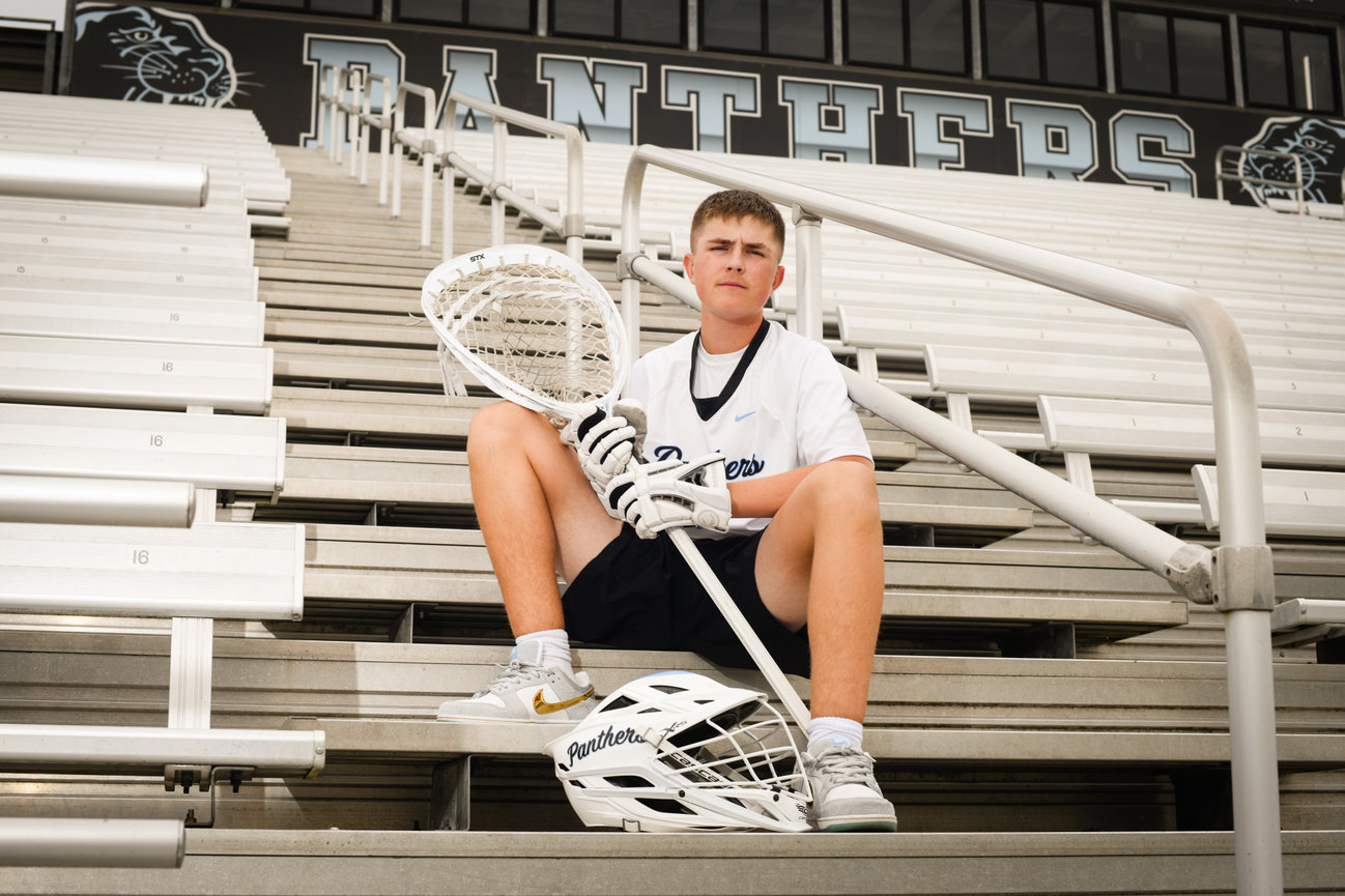 Lacrosse athlete sitting on bleachers, holding a stick and wearing a team jersey.