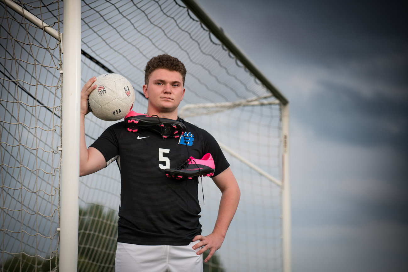 Teenage soccer player in black jersey poses with a soccer ball and pink cleats near a goalpost.