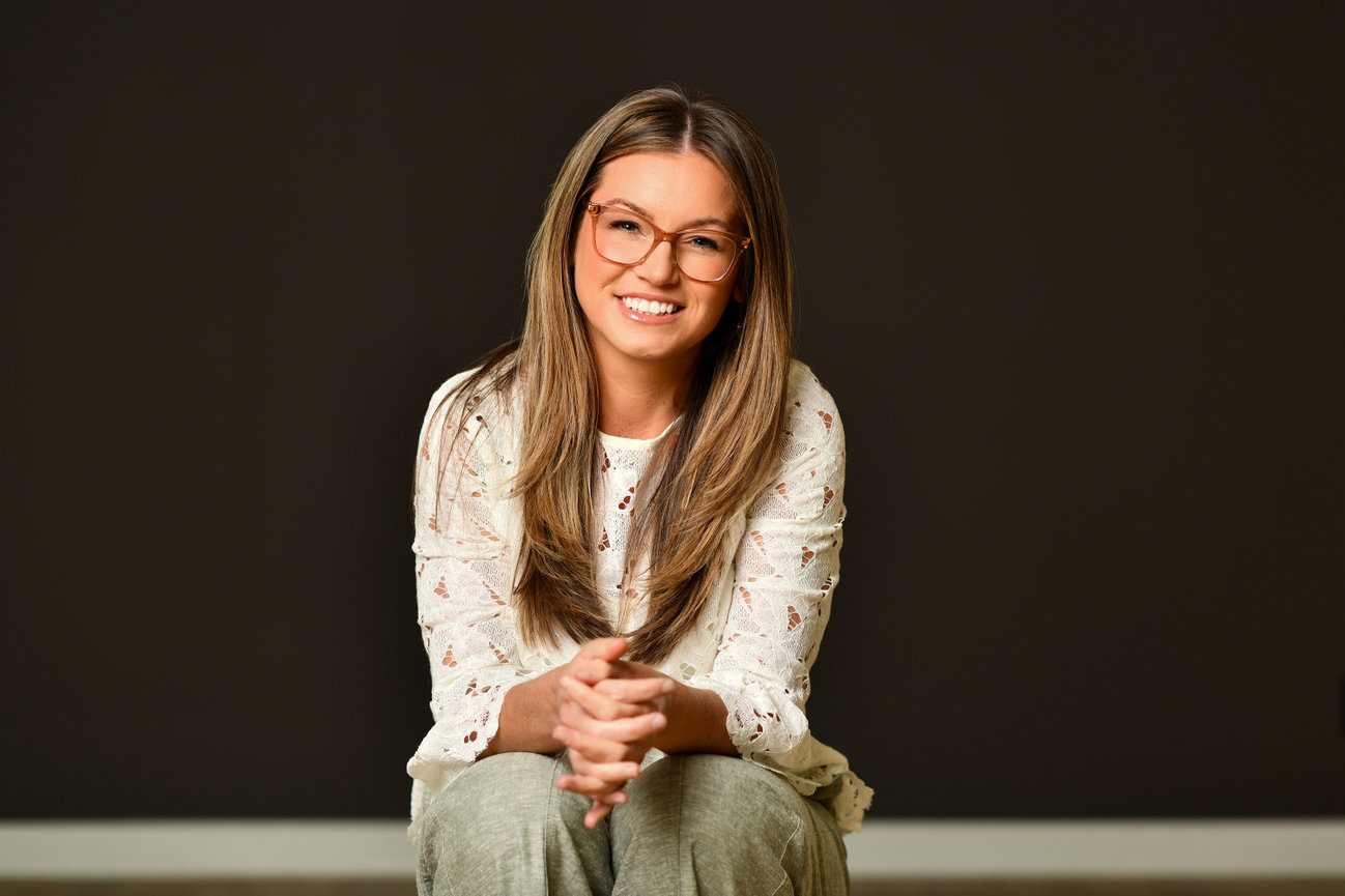 Smiling young woman with long hair and glasses, seated against a dark background, wearing a white top and light pants. Therapist being photographed for updated headshots and professional branding in dublin ohio.