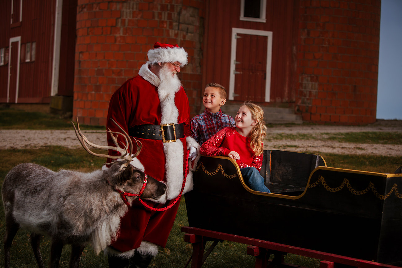 At a previous Kay Web Photography experience, Santa talks with smiling children beside a reindeer and sleigh